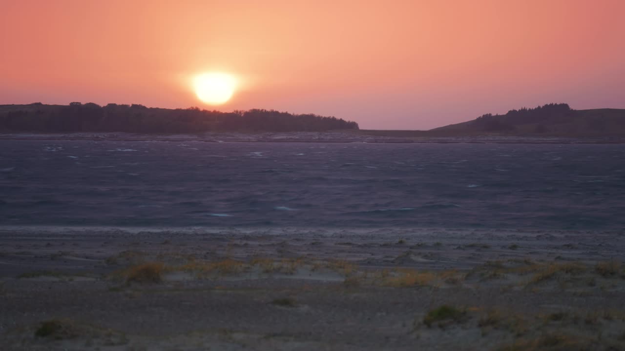 espectacular espectáculo de una puesta de sol roja pintando el cielo sobre el fiordo, proyectando un resplandor ardiente sobre la playa de arena de abajo, mientras las olas ruedan lentamente sobre la arena en un video timelapse