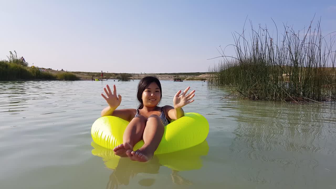 Girl sitting in yellow inflatable tire floating in lake, looking and waving at camera during beautiful summer day