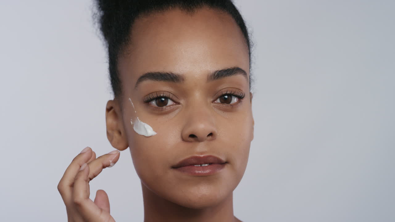 retrato hermosa joven mujer afroamericana aplicando crema hidratante para la piel en la cara disfrutando de exfoliación masajeando loción antienvejecimiento de lujo belleza femenina perfecta en fondo blanco concepto de cuidado de la piel