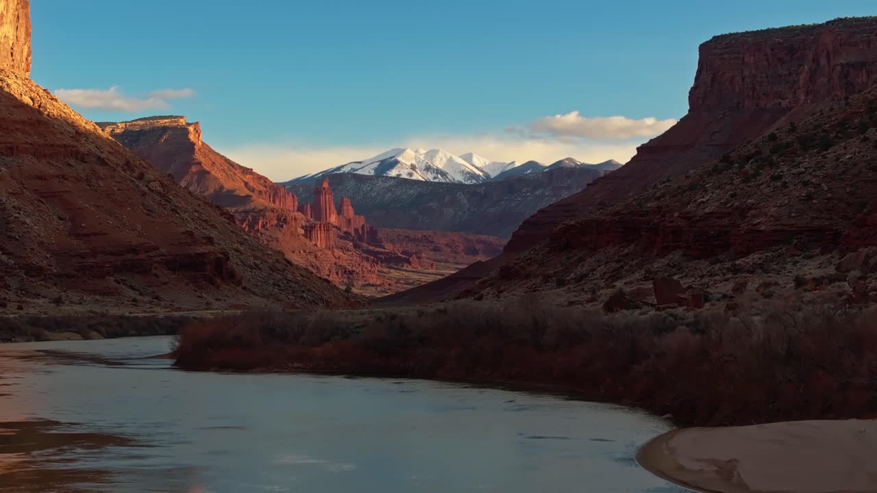 Sunset glows across calm desert river canyon with mirrored reflections and warm sandstone walls, snowcapped mountain in distance, Moab Utah USA