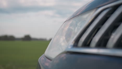 close up of modern car headlamp blinking beside quiet countryside road, soft reflections on chrome edges, distant green field blurred in background beneath pale sky with gentle clouds