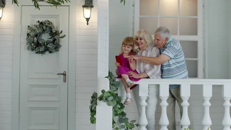 Senior-ouple-sitting-with-granddaughter-in-porch-at-home.-Using-mobile-phone-for-online-video-call