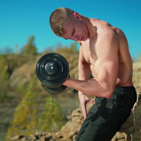 muscular man is lifting metal dumbbell in nature. shirtless sportsman exercises shoulder muscles near the rock against blue sky.