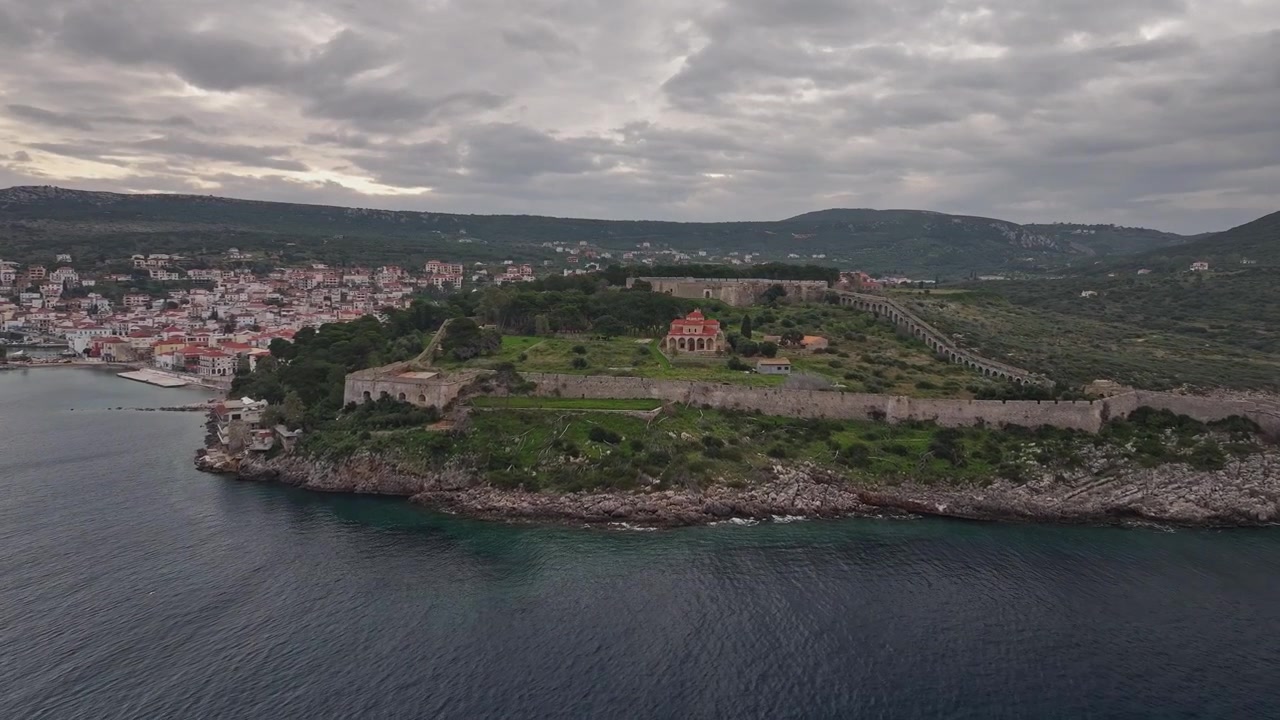 Ripresa aerea con drone in avanti che avanza verso il promontorio della fortezza di Niokastro, mostrando le sue mura in pietra, la Chiesa della Trasfigurazione, l'acquedotto ottomano e la città di Pylos sotto un cielo nuvoloso al tramonto in Grecia.