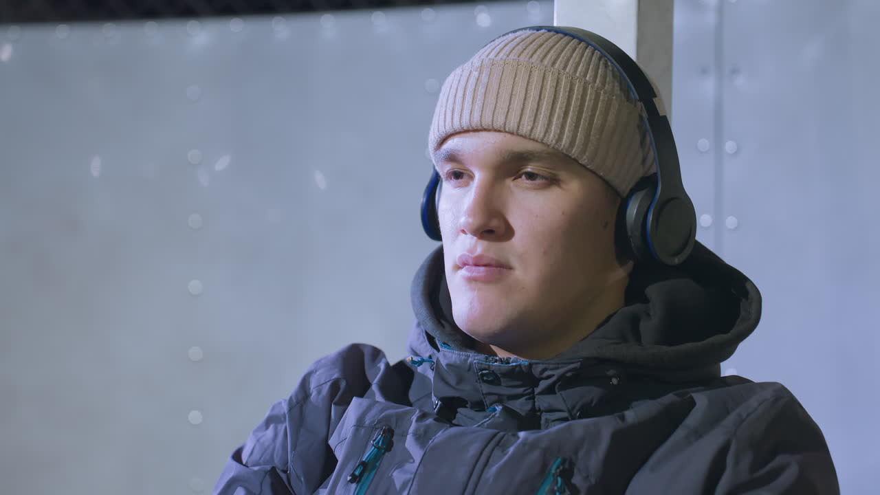 primer plano de un joven pensativo con gorra y auriculares contra un telón de fondo deportivo neutral, su expresión reflexiva, profundamente en pensamiento con un entorno deportivo alrededor