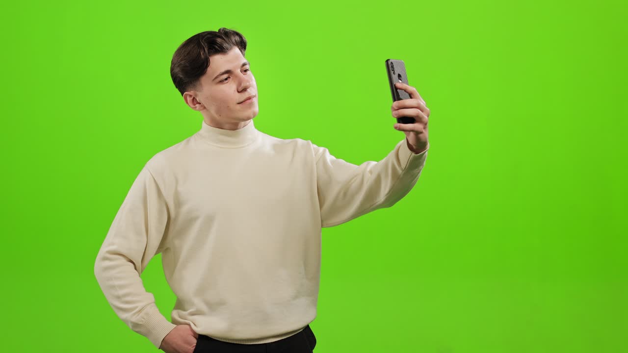 한 남자가 스마트폰으로 셀피를 찍고, 주머니에 손을 넣고, 초록색 바탕에 서 있다.
