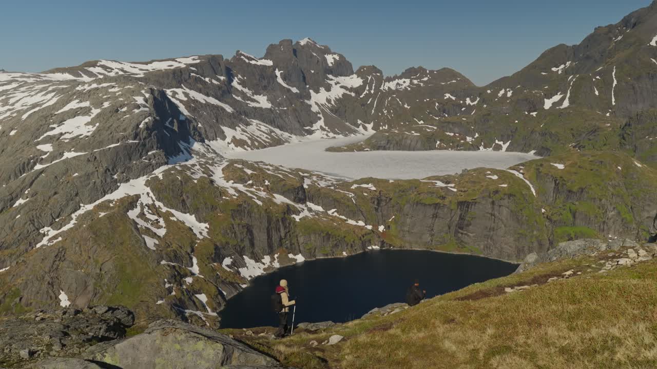 Turisté sestupují k úchvatnému alpskému jezeru s výhledem na výše položené zamrzlé jezero a zasněžené žulové hory pod jasně modrou oblohou na Lofotech v Norsku, zachycující dramatickou horskou krajinu.