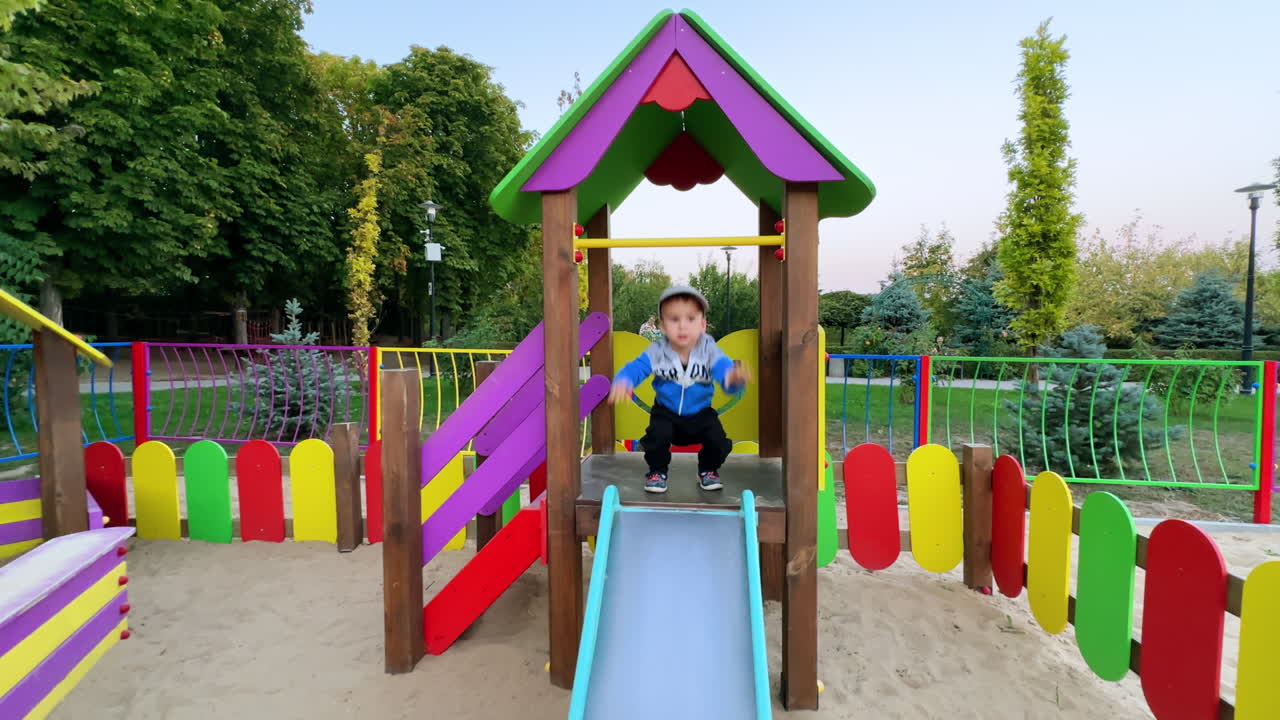 Cute kid in a cap hanging on the horizontal bar. Happy kid hitting his feet by the slide and then goes down.