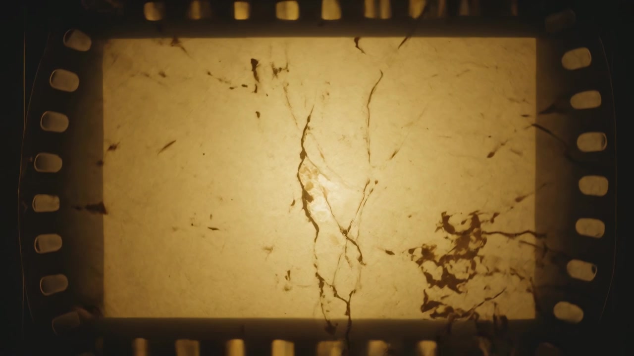 Tira de película de 35mm avanzando sobre los piñones, revelando grietas, polvo y arañazos en una lámpara ámbar para escanear