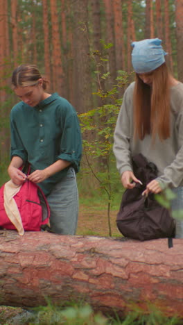 los excursionistas se acercan al árbol caído en el bosque, cada uno con mochilas en la mano, una con bandana azul lucha por abrir su bolsa, mientras que la otra coloca la suya en el tronco, ambos centrados en su tarea en medio de árboles altos