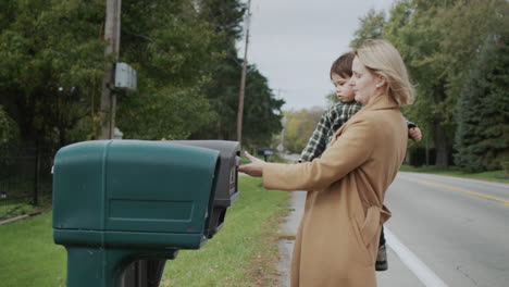 a woman with a child in her arms picks up a letter from a street mailbox