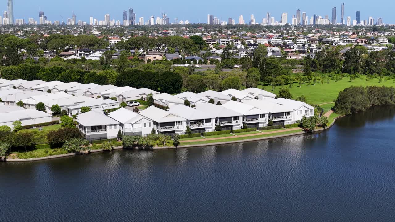 Drone pans across modern lakeside homes toward distant Gold Coast skyline under bright daylight