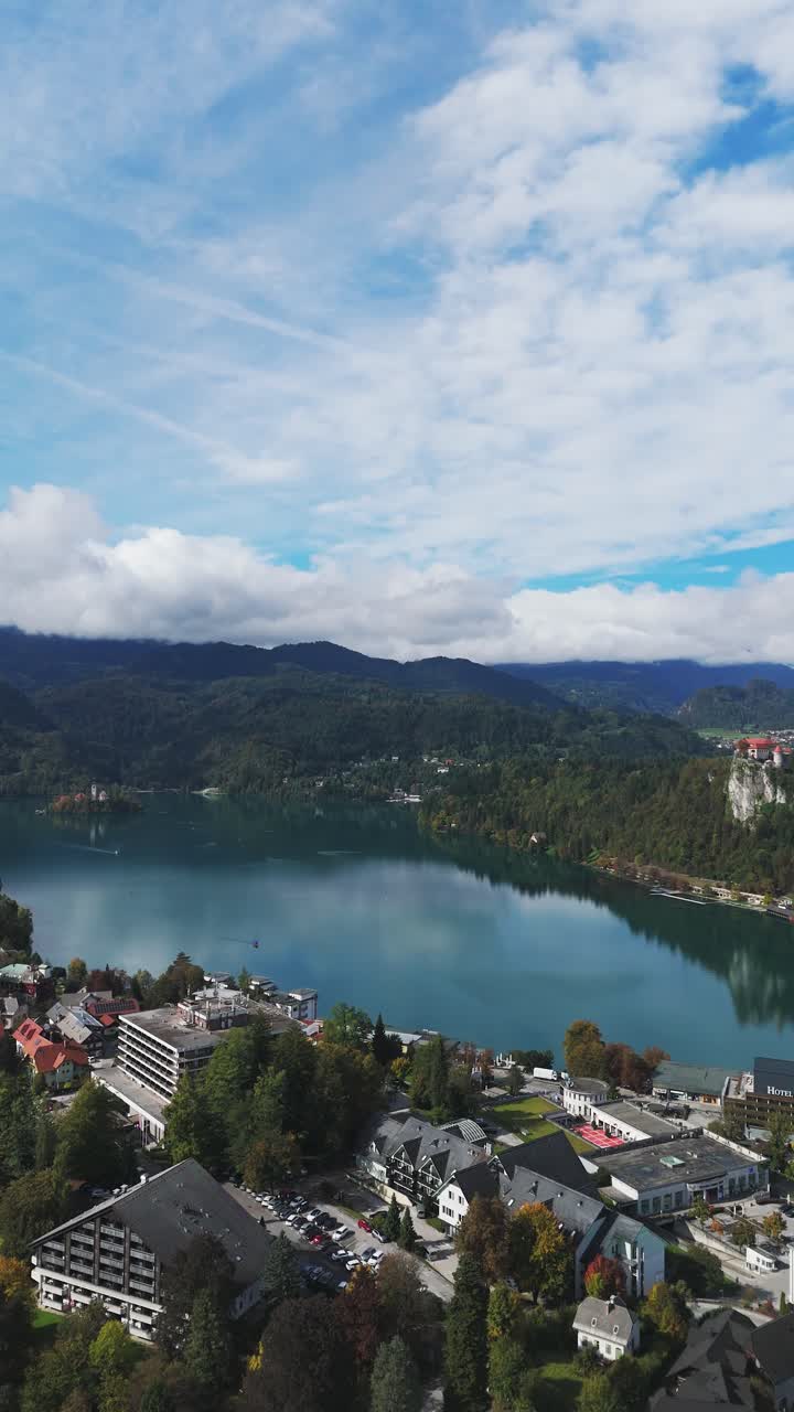 Lenta ripresa verticale con drone dell'iconico lago di Bled, Slovenia: acqua cristallina, isola con chiesa, castello arroccato in alto, circondato da foreste autunnali e montagne suggestive