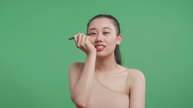 Asiatische Frau, die Eyeliner aufträgt, perfekte Lidstriche zieht und lächelt, während sie vor einem Greenscreen-Hintergrund im Studio steht.