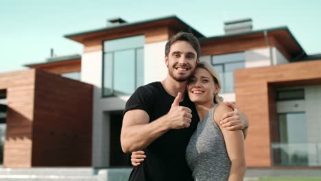 pareja feliz haciendo pulgares arriba cerca de una casa de lujo. familia emocionada sonriendo afuera