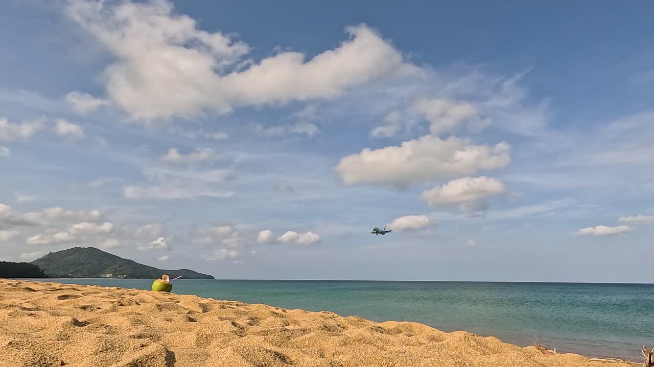 A plane approaches and lands over a sunny beach in Phuket, Thailand, with clear skies and a serene ocean backdrop