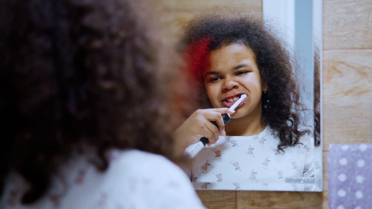 Cute african american child diligently brushing her teeth using electric toothbrush, looking at reflection in bathroom mirror, focused on maintaining good oral hygiene