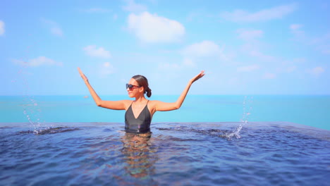 happy asian woman in black bathing suit and red sunglasses inside rooftop infinity pool water splashing water with hands, cloudy sky and sea on background