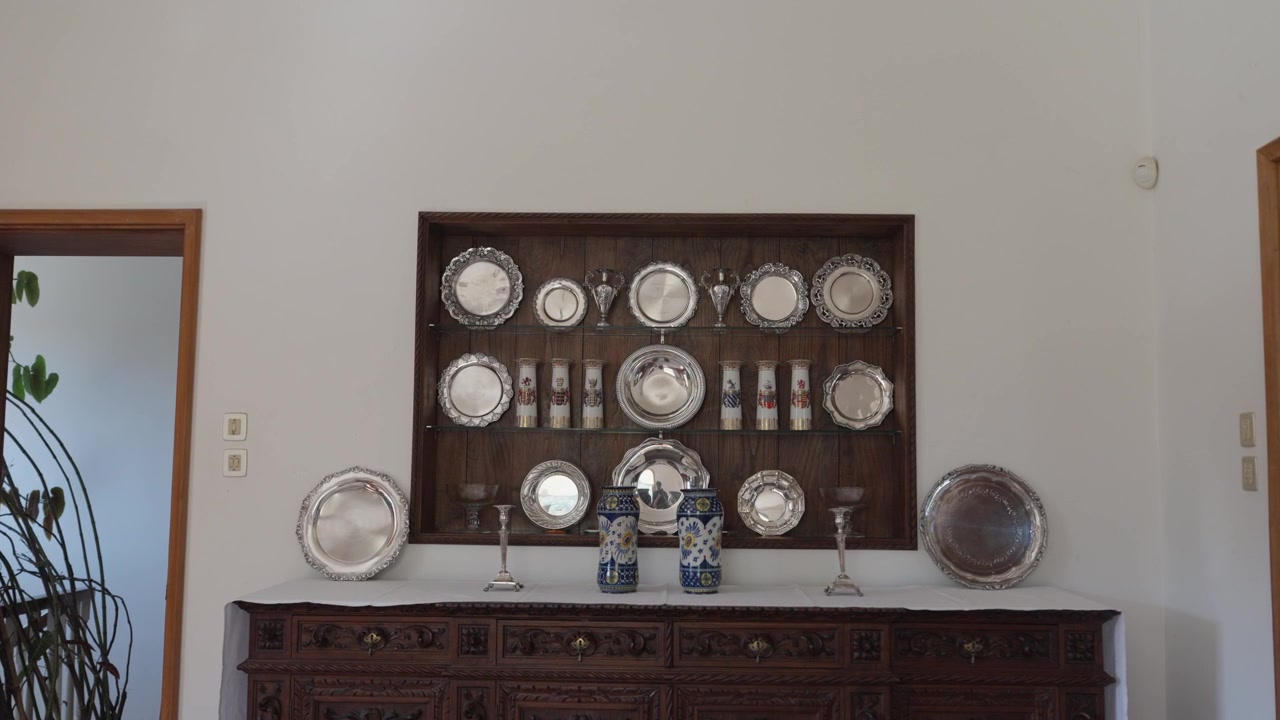 Antique silver platters displayed in a wall niche above a carved wooden sideboard, with blue and white vases in a bright room.