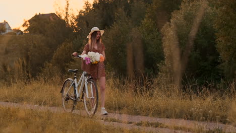 Young-beautiful-blonde-girl-in-summer-in-dress-and-hat-walking-on-road-with-bike-and-flowers-in-slow-motion.