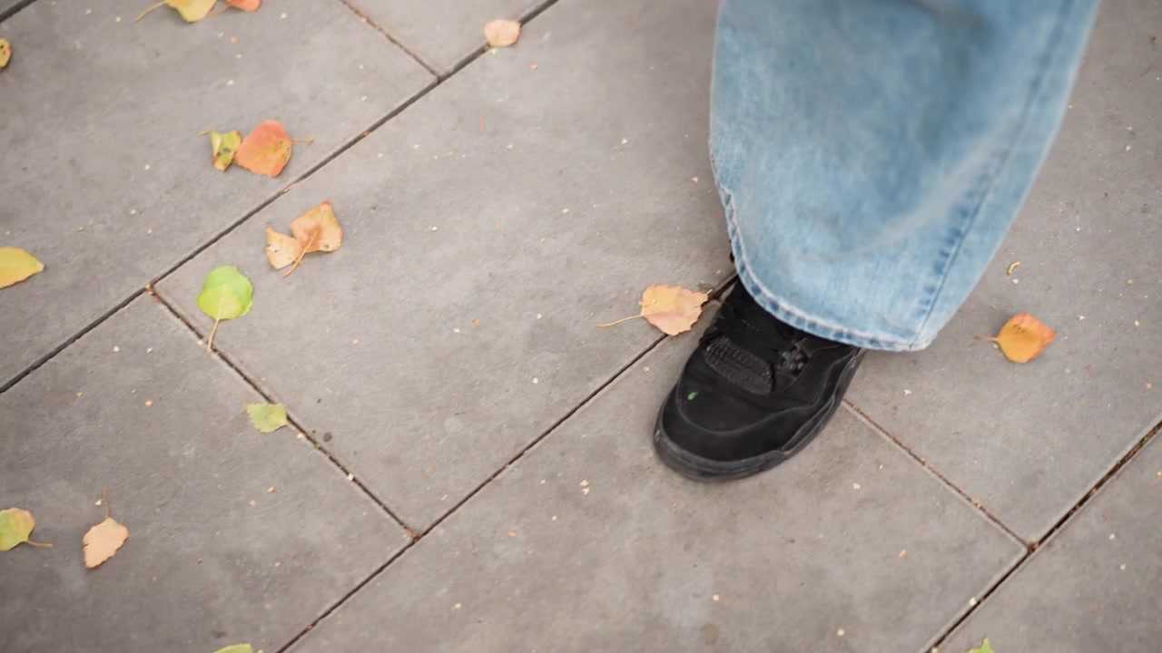 Close up of person feet in black sneakers and blue jeans walking on gray stone pavement with scattered autumn leaves. scene captures movement through outdoor environment focusing solely