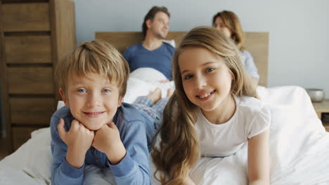 cute small boy and her sister lying and smiling at camera, his parents are lying under the blanket on the bed behind him