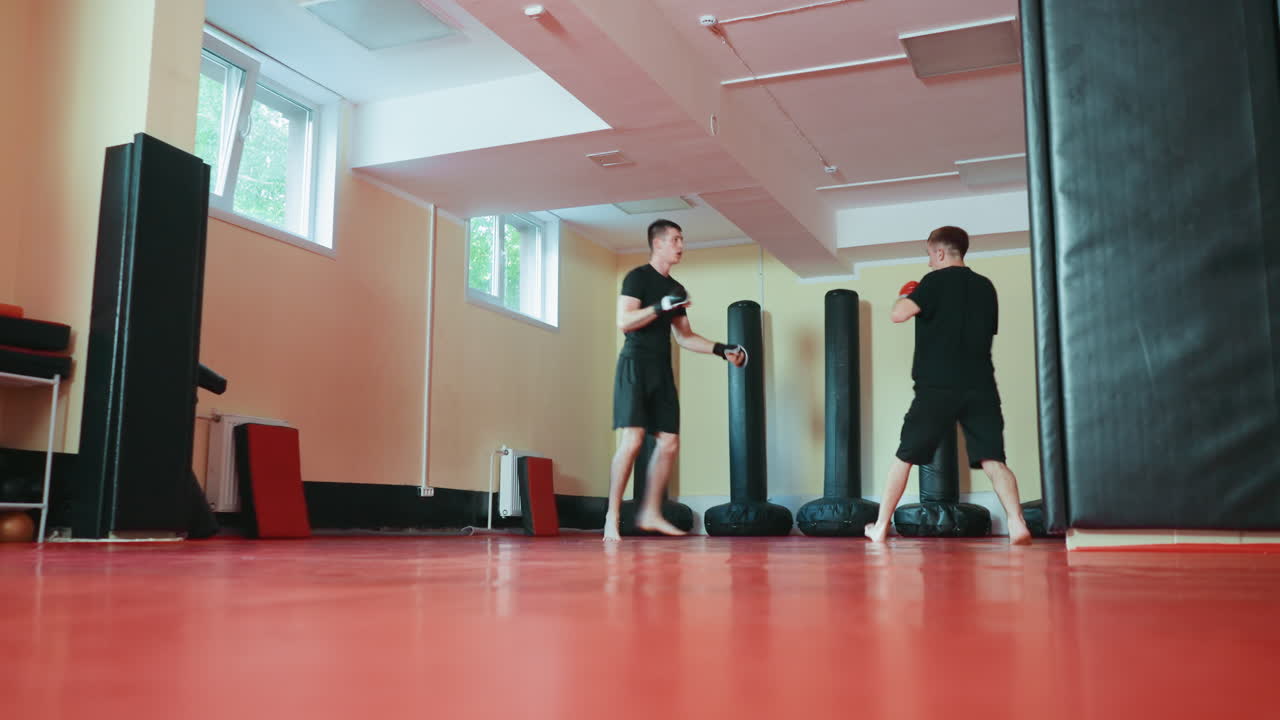 Two boxers sparring in martial arts gym as one lifts knee preparing for kick while opponent guards with gloves on red mat floor surrounded by punching bags during combat training session under daylight