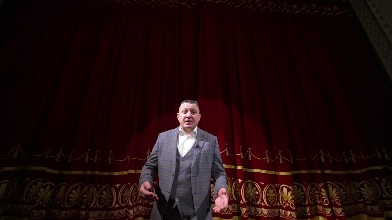 Man in gray costume on stage. Middle-aged actor talking on camera on luxury red curtains background in the theater. Slow motion.