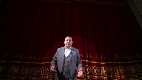 man in gray costume on stage. middle-aged actor talking on camera on luxury red curtains background in the theater. slow motion.