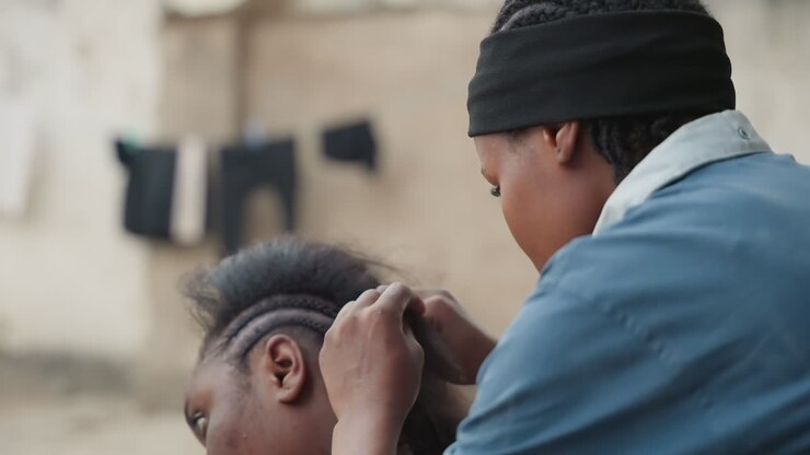 Outdoor closeup braiding hair gently. Home stylist in beanie carefully parting scalp while plaiting tight rows, hands focused, neighbor seated, laundry on wall in background, soft daylight, intimate