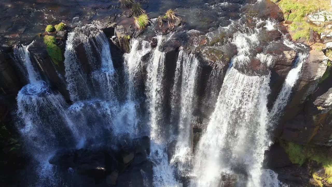 Drone footage captures Ebor Falls in New South Wales, Australia, with multiple streams of water plunging over rugged cliffs in bright daylight