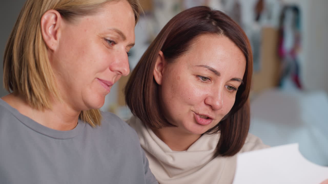 Women smiling warmly while concentrating on project selection, analyzing design details together in bright workspace, surrounded by colorful fabric samples, sharing ideas with focus and enthusiasm