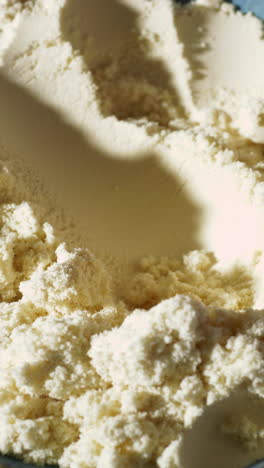 a black scoop presses into a fluffy, pale yellow protein powder, possibly vanilla or banana flavored, creating a soft texture detail in this close-up shot under warm, directional light.