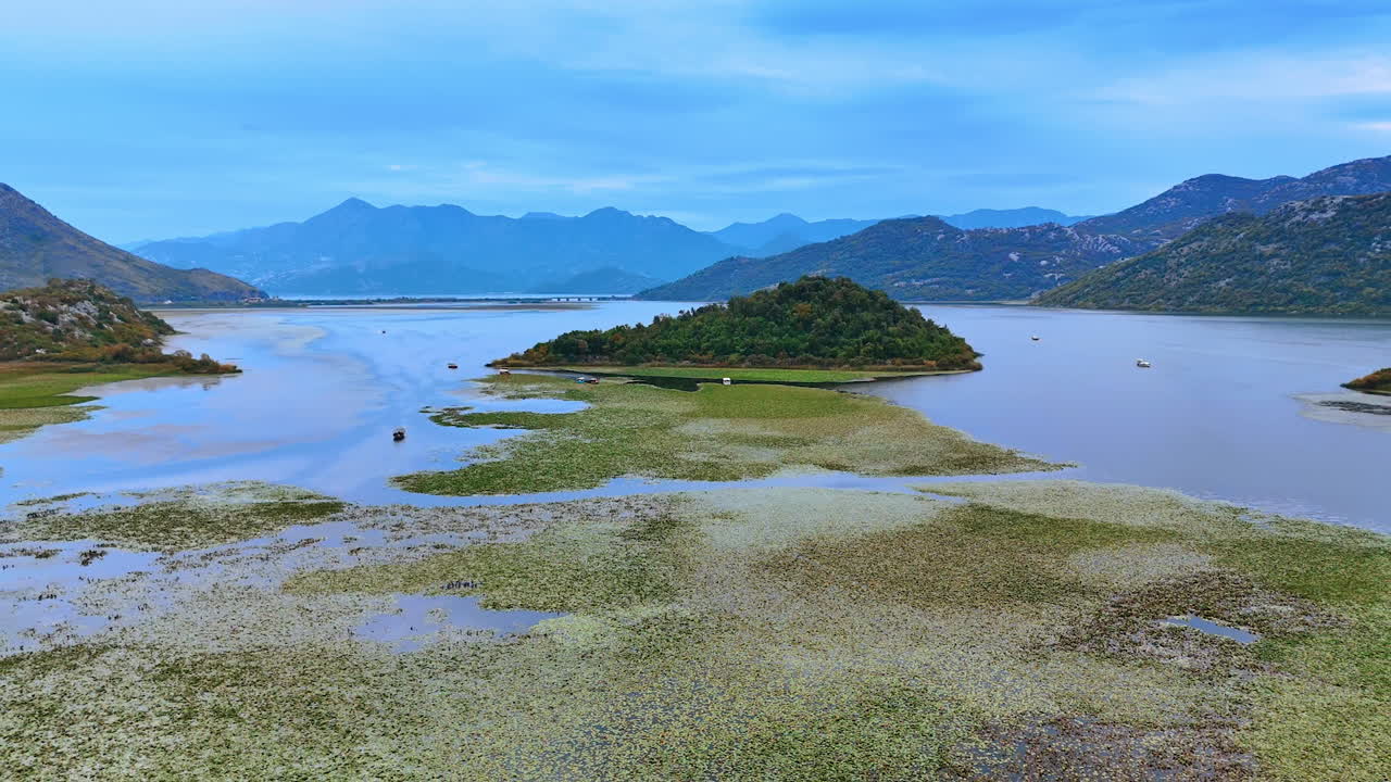 Distancing from the little rocky island covered with vegetation. Multiple water lily pads grow on the surface of waterscape. Lake Skadar, Montenegro and Albania border. Stunning mountains at backdrop