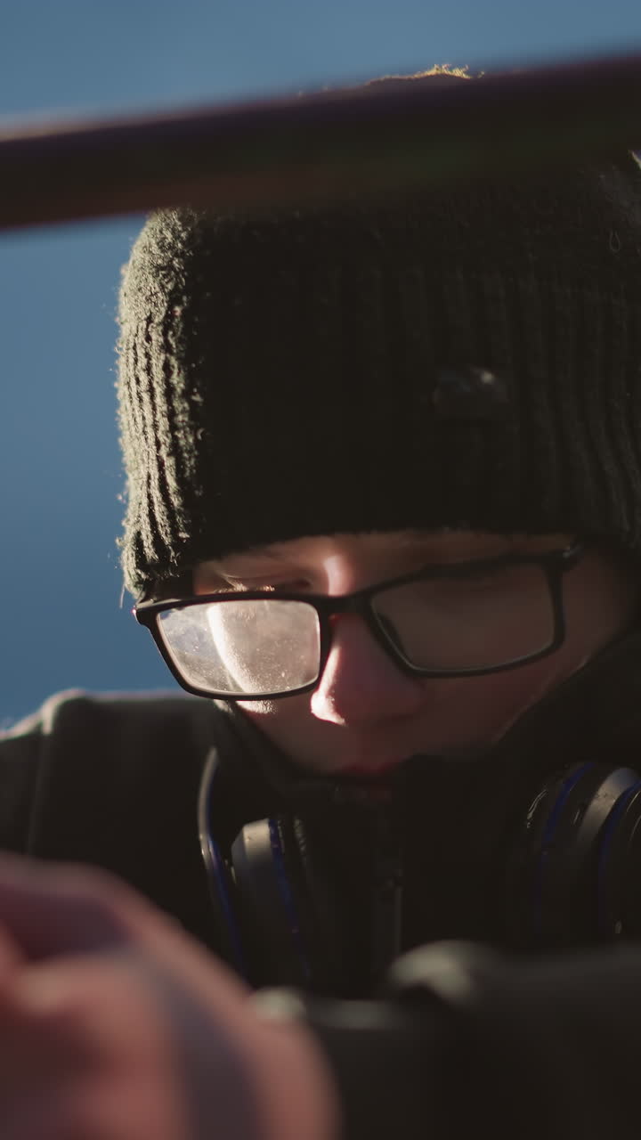 Close-up of a young boy wearing glasses and a black jacket, with his head bowed and looking thoughtful, he has a headset around his neck, and his hands are clasped in front of him