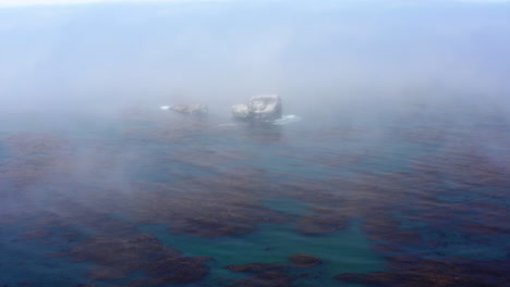 huge rocks sticking out of the sea, barely seen through the mist that flows over the water