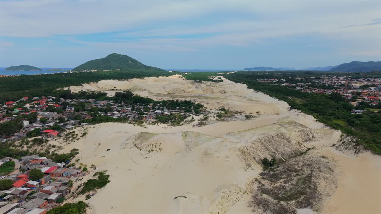Ripresa aerea con tracciamento a destra sulle Dune di Santinho (Dunas do Santinho). Dune di sabbia bianca sono intervallate da case residenziali e vegetazione costiera vicino all'oceano