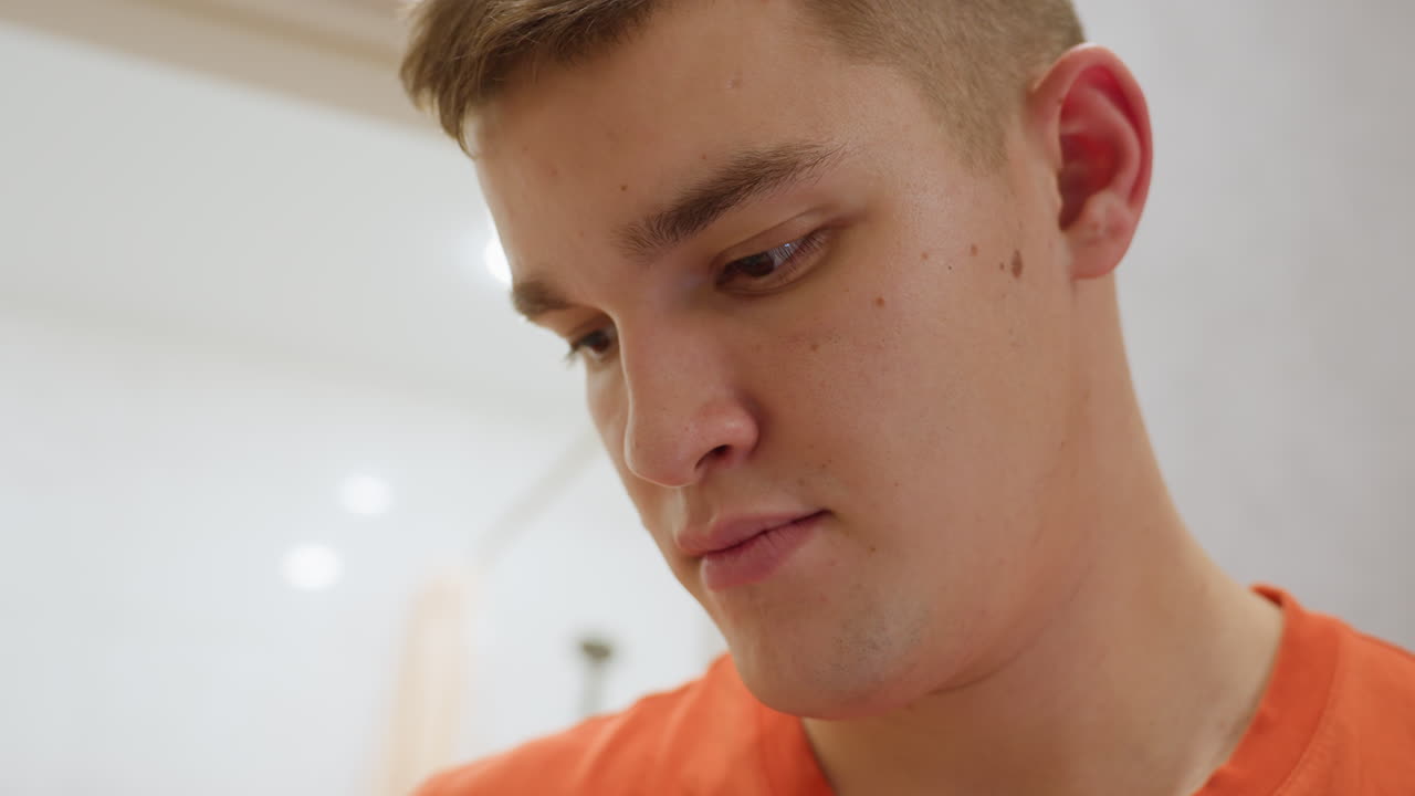 Close up of man in orange shirt looking attentively at something in background with focused expression under bright indoor lighting showing detail of face concentration