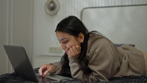 Ein-Glückliches,-Braunhäutiges,-Brünettes-Mädchen-In-Einem-Grauen-Pullover-Liegt-Auf-Dem-Bett-Und-Tippt-Auf-Ihrem-Laptop,-Während-Sie-In-Einem-Modernen-Apartment-Im-Schlafzimmer-Arbeitet-Und-Zeit-Online-Verbringt