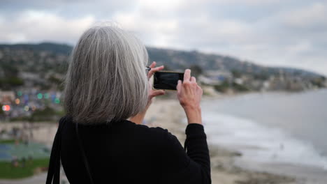 A-middle-aged-woman-on-vacation-taking-a-picture-with-her-phone-of-the-city-and-ocean-in-Laguna-Beach,-California-SLOW-MOTION