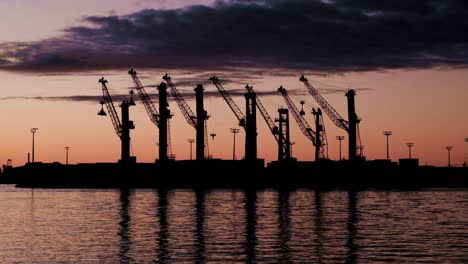 Silhouettes-of-huge-maritime-shipping-container-cranes-at-sunset-in-a-harbour