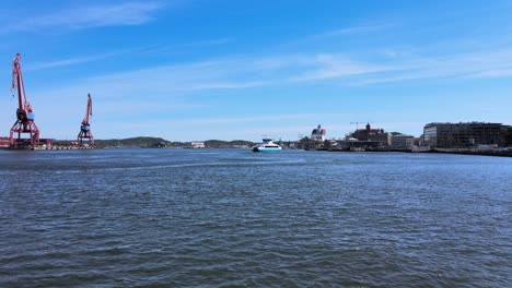 forward areial drone flight towards a small passanger ferry that is about to arrive to stenpiren in gothenburg, sweden on a beautiful day with the ship yard cranes and city scape in the backgrund