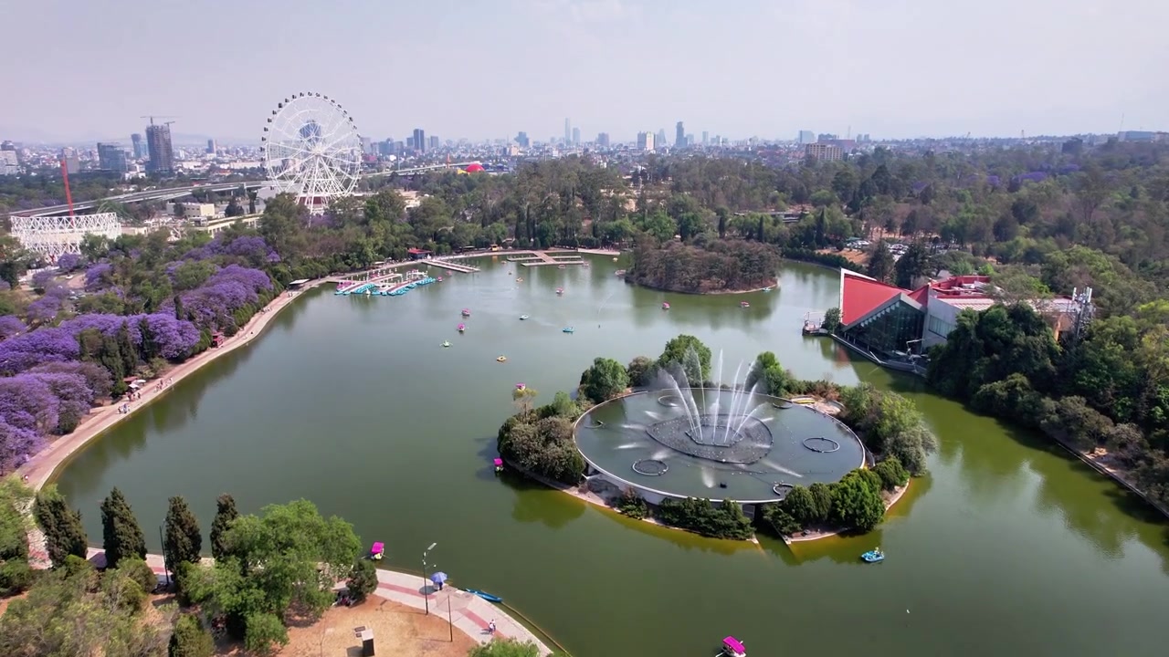 Voo de drone sobre o lago da Segunda Seção de Chapultepec, destacando a fonte central, a roda-gigante Aztlán, as árvores de jacarandá e o horizonte da Cidade do México em um dia ensolarado