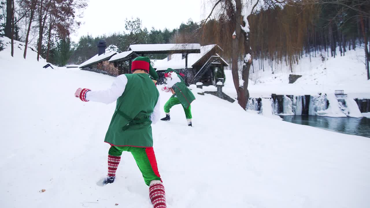 Happy elves in green fairy costumes outdoors in winter. Two cute elves playing snowballs near the river on forest background