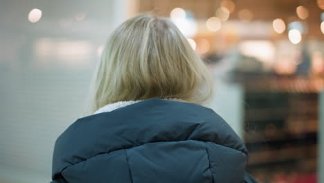 안경과 검은색 후디를 입은 젊은 여성이 잘 조명된 쇼핑몰을 고 있는 뒤쪽 헤드 뷰, 흐릿한 배경을 가진 반사와 함께 유리를 통해 볼 수 있는 부드러운 보케 빛 효과