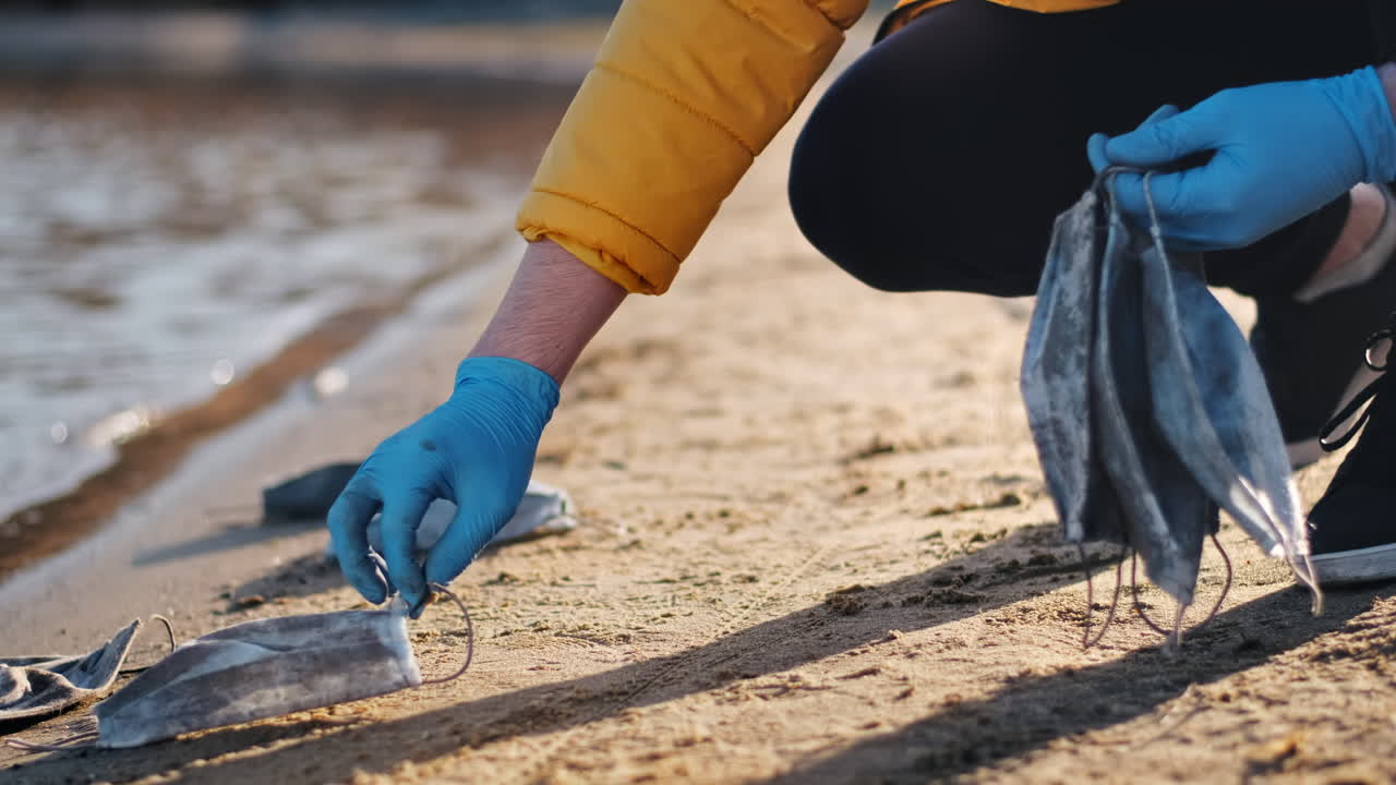 Man in medical gloves picking up a bunch of dirty medical masks from the beach of a lake. Pollution idea
