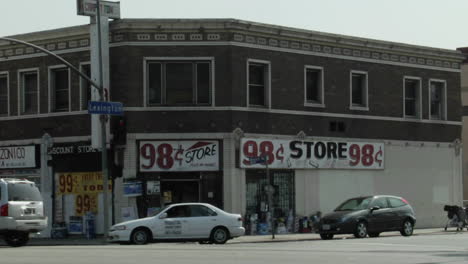 traffic moves through a city street as pedestrians walk by with a 98 cent store background