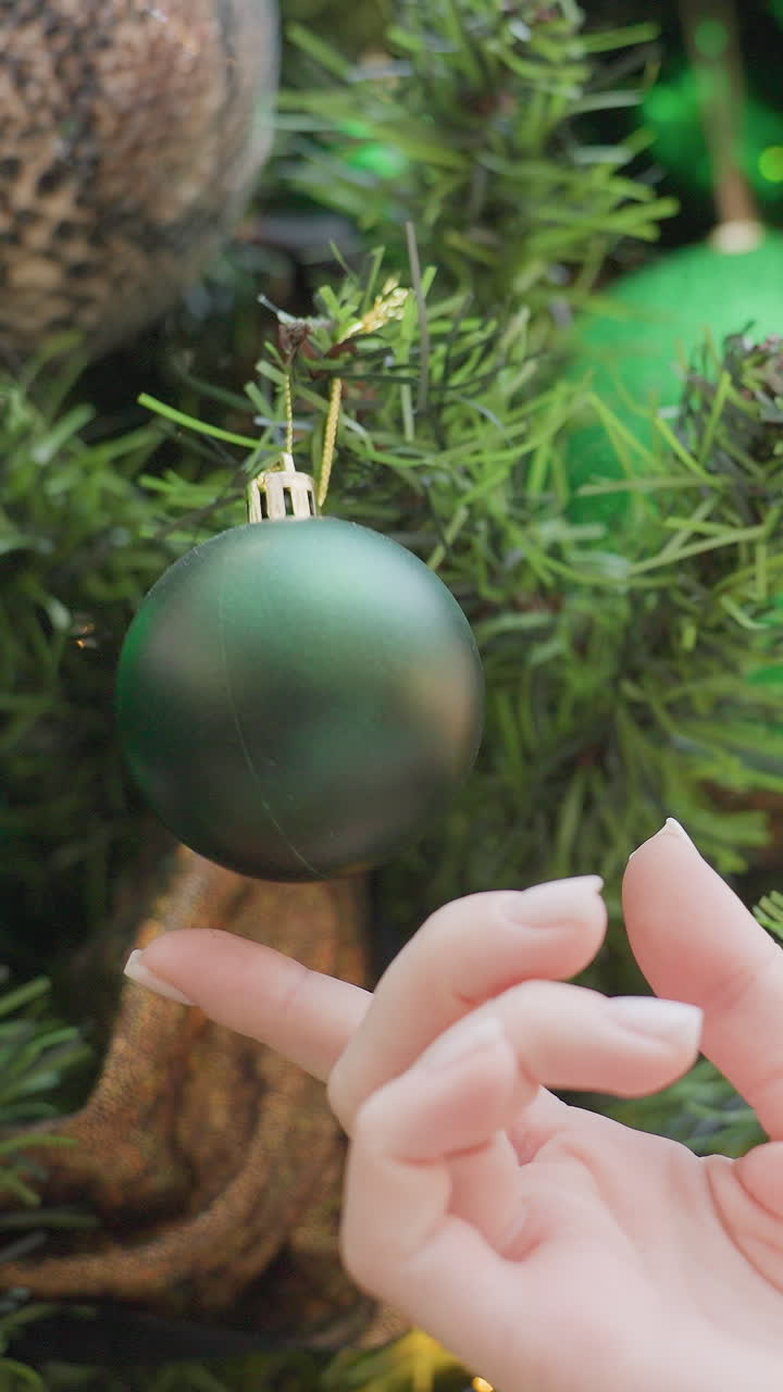 Close-up of hand holding small green decorative Christmas ball from tree, with lush greenery and soft, warm decorative lights shining