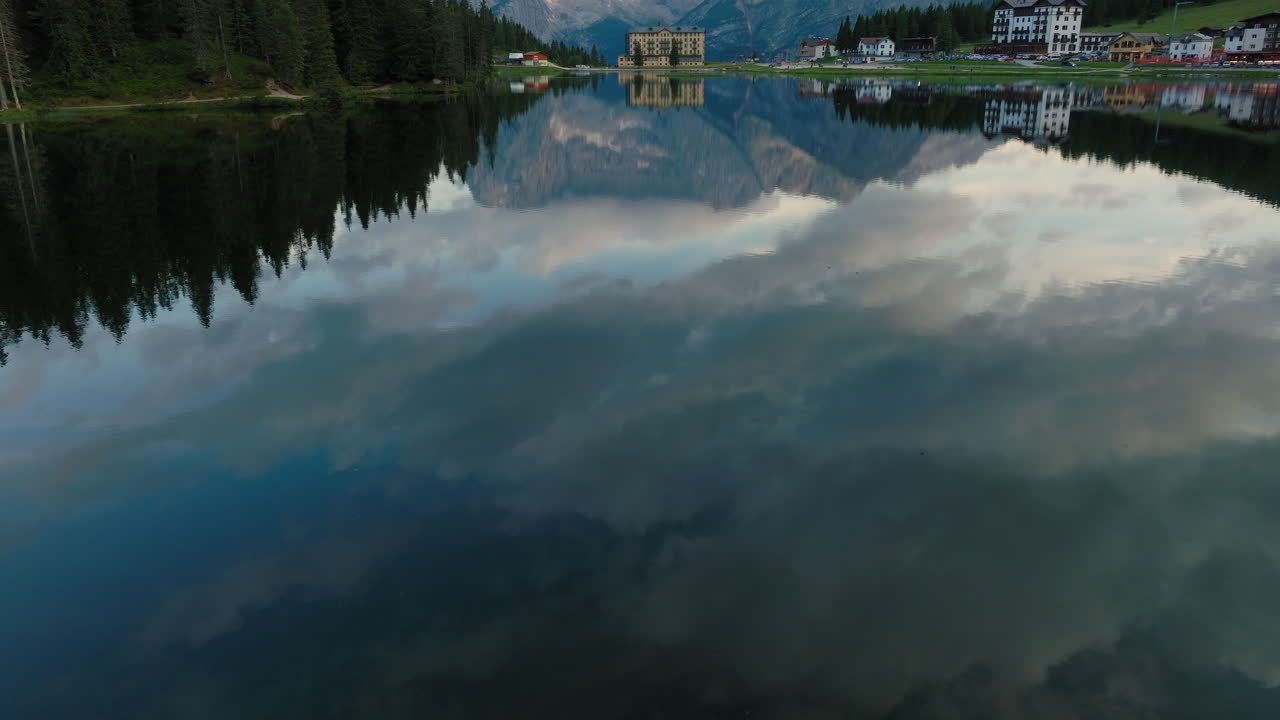 Slow drone reveal shot of Lake Misurina during spring with calm, mirror-like waters reflecting the surrounding alpine peaks and forests