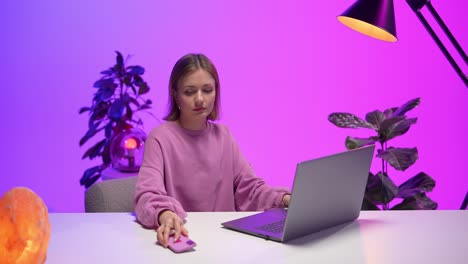 young woman in a pink sweatshirt multitasking at a sleek white desk, holding a smartphone while using her laptop, set in a vibrant workspace with purple lighting and lush green houseplants